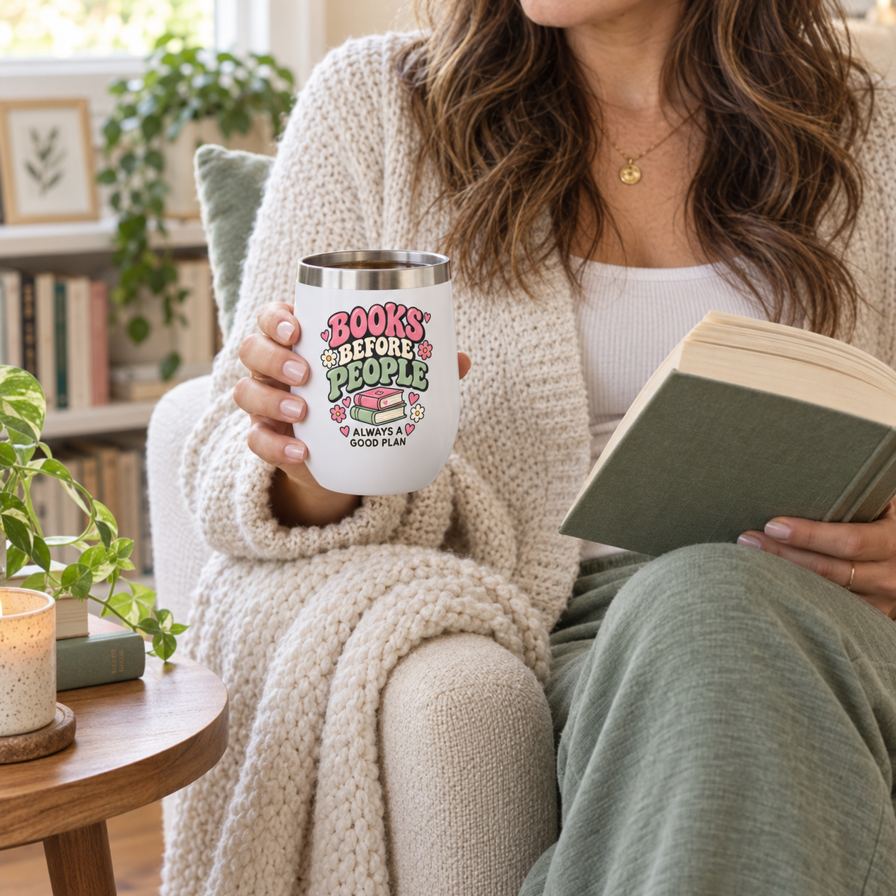 A woman in a cozy living room setting holds a white insulated tumbler with a stainless steel rim. The tumbler features colorful text that reads 'BOOKS BEFORE PEOPLE IS ALWAYS A GOOD PLAN' with a pink book illustration.