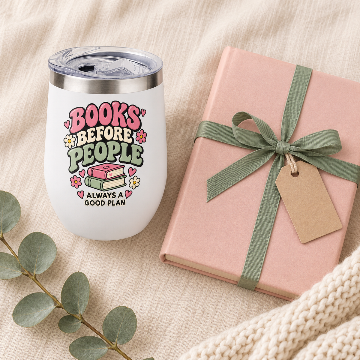 A white insulated tumbler with a silver lid and a colorful book-themed graphic reading 'BOOKS BEFORE PEOPLE' and 'ALWAYS A GOOD PLAN,' styled on a beige fabric surface beside a pink gift box tied with a green ribbon and a small kraft tag, with eucalyptus leaves and a cream knit blanket nearby.