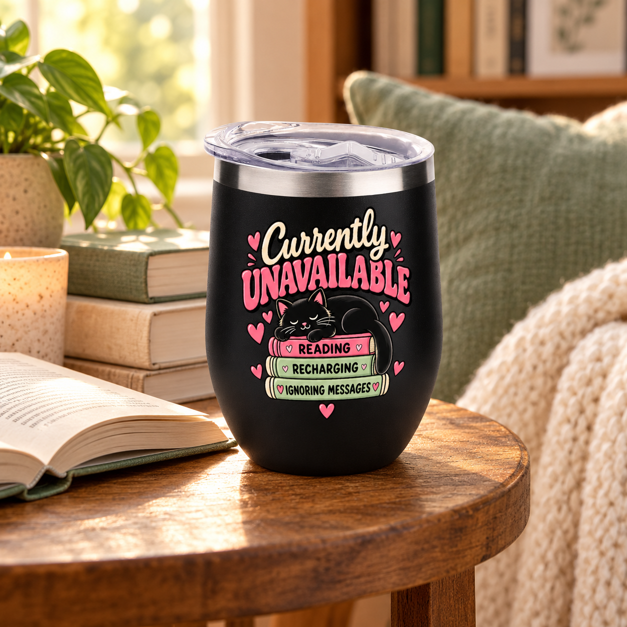 A black insulated tumbler with a clear lid sits on a wooden table in a cozy indoor setting. The tumbler has a printed design featuring the phrase 'Currently UNAVAILABLE' in pink and white, a sleeping black cat, and pink hearts.