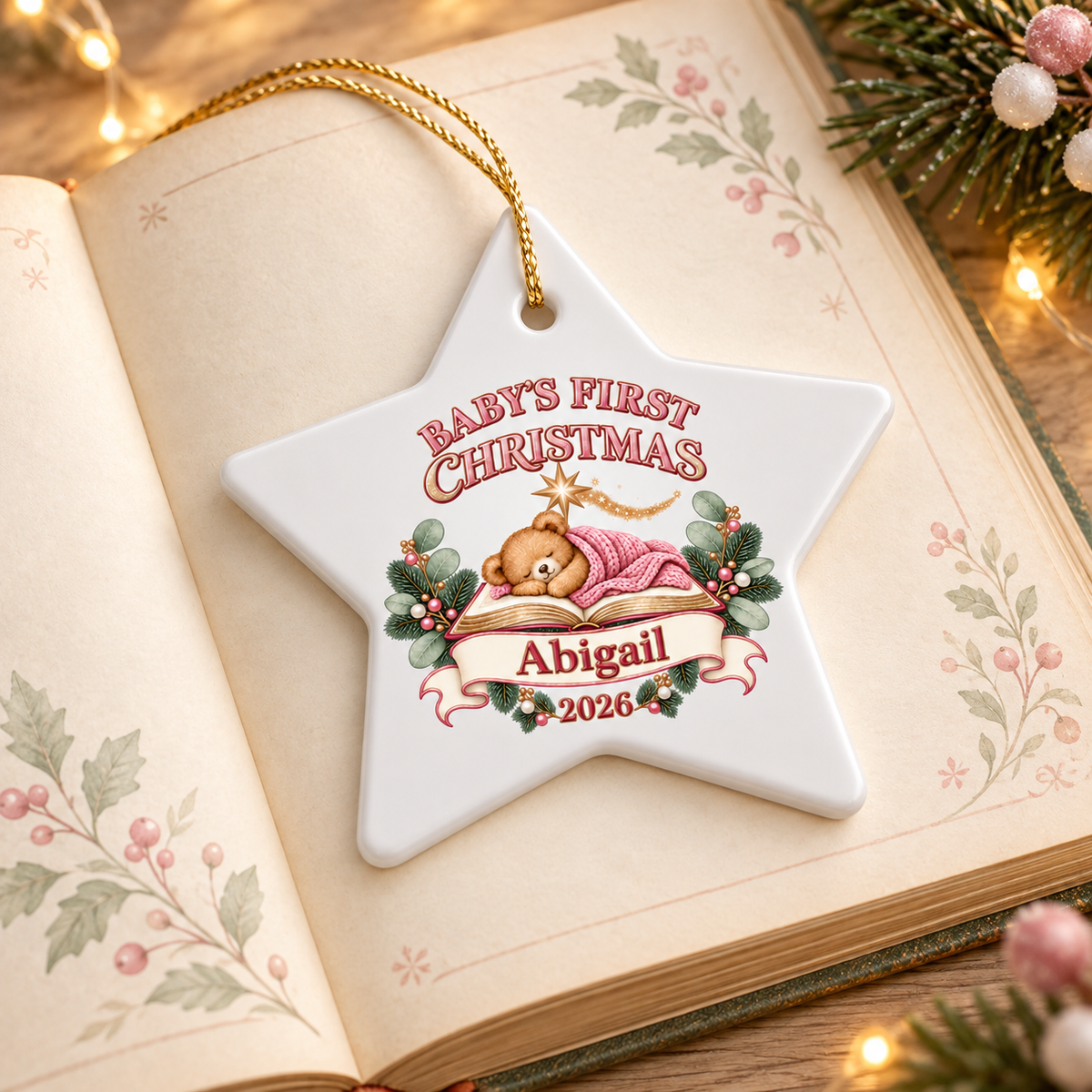 A personalized white star-shaped Christmas ornament with a gold hanging string, featuring an illustration of a teddy bear and the text 'Baby's First Christmas', 'Abigail', and '2026'. The ornament is styled on an open vintage book with warm fairy lights and pine branches in the background.