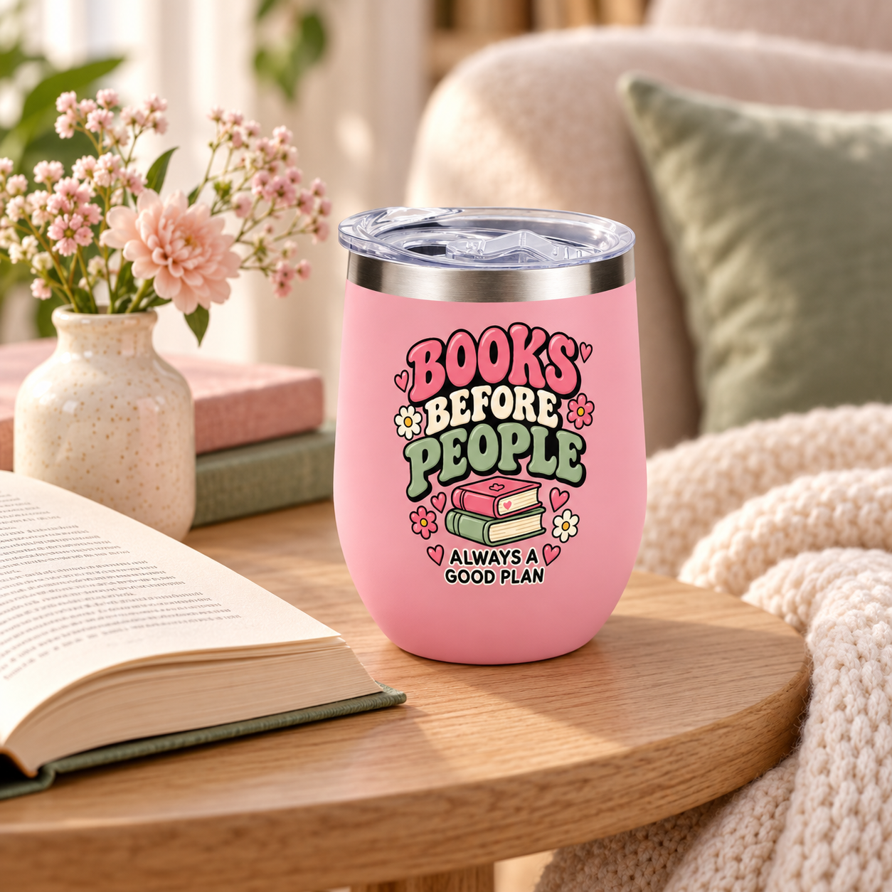 A pink insulated stemless tumbler with a clear lid sits on a wooden table in a cozy living-room setting. The tumbler is printed with the phrase “BOOKS BEFORE PEOPLE” and “ALWAYS A GOOD PLAN” with illustrations of books, flowers, and hearts.