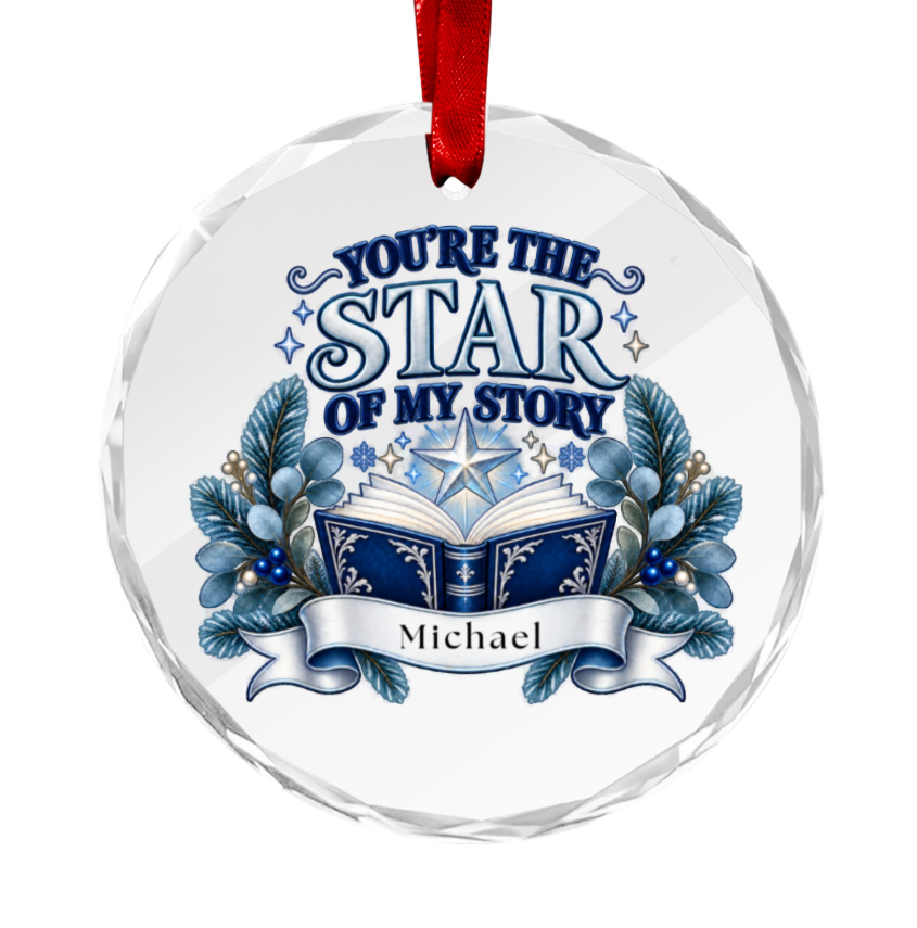 A round clear hanging ornament with a red ribbon loop at the top. The ornament features printed blue and white decorative text reading “YOU’RE THE STAR OF MY STORY” above an illustrated open book, blue berries, and a banner reading “Michael.