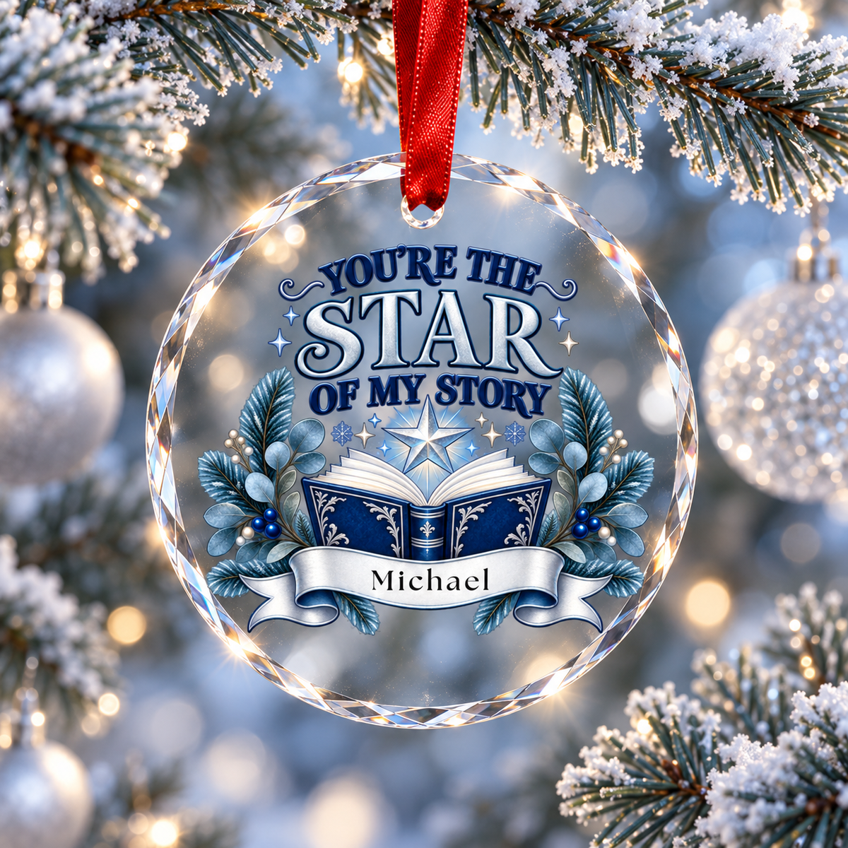 A personalized clear glass Christmas ornament hanging from a red ribbon, featuring the text 'YOU'RE THE STAR OF MY STORY' above an open book and blue evergreen branches, with the name 'Michael' on a banner. The ornament is set against a festive blurred Christmas tree background with snow-dusted branches and silver ornaments.