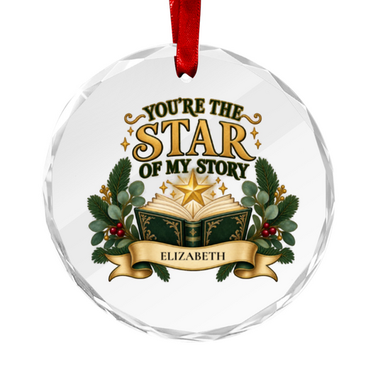 A round clear faceted glass or acrylic ornament hanging from a red ribbon. The ornament features printed text reading 'YOU'RE THE STAR OF MY STORY' above an open book with a glowing star, green holly leaves, red berries, and a gold banner reading 'ELIZABETH'.