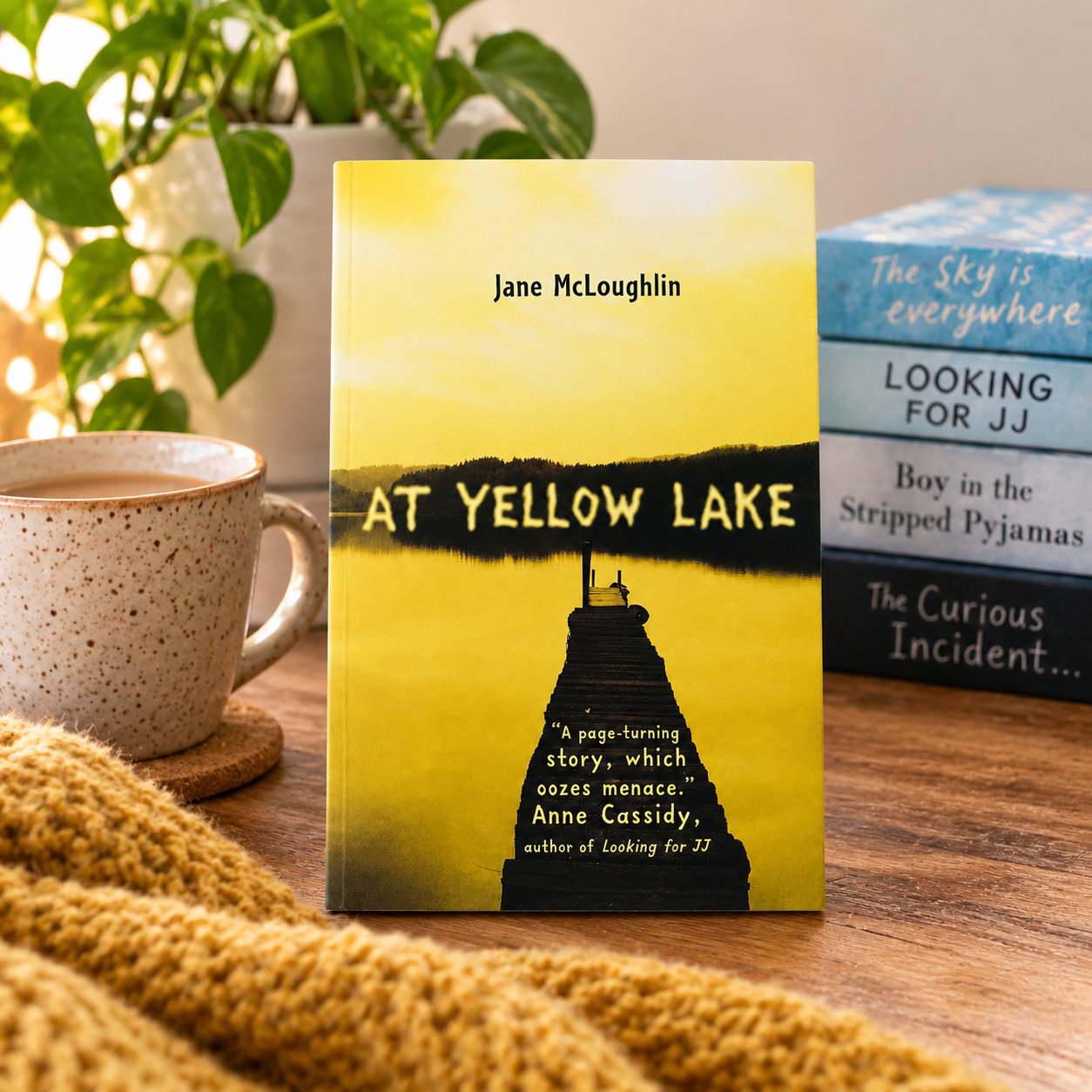 A book titled "At Yellow Lake" by Jane McLoughlin stands upright on a wooden table, surrounded by a mug, a knitted blanket, and other books in the background. The book cover is predominantly yellow with a black silhouette of a lighthouse and bold black title text.