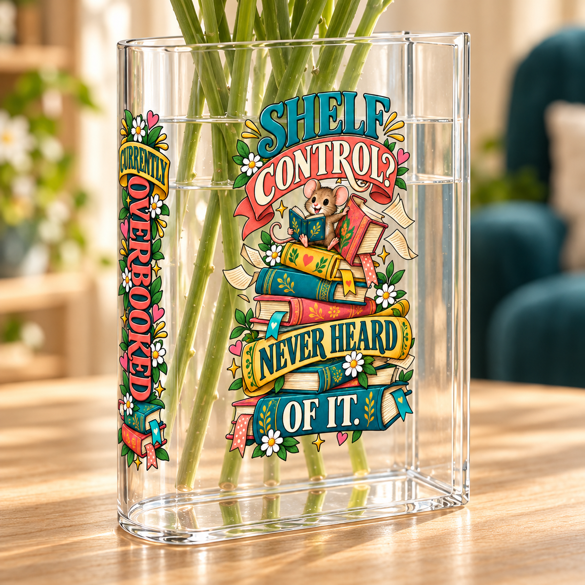 A clear glass tumbler with a colorful book-themed graphic that reads 'SHELF CONTROL? NEVER HEARD OF IT.' and 'OVERBOOKED,' shown filled with green stems on a wooden table in a softly blurred indoor setting.