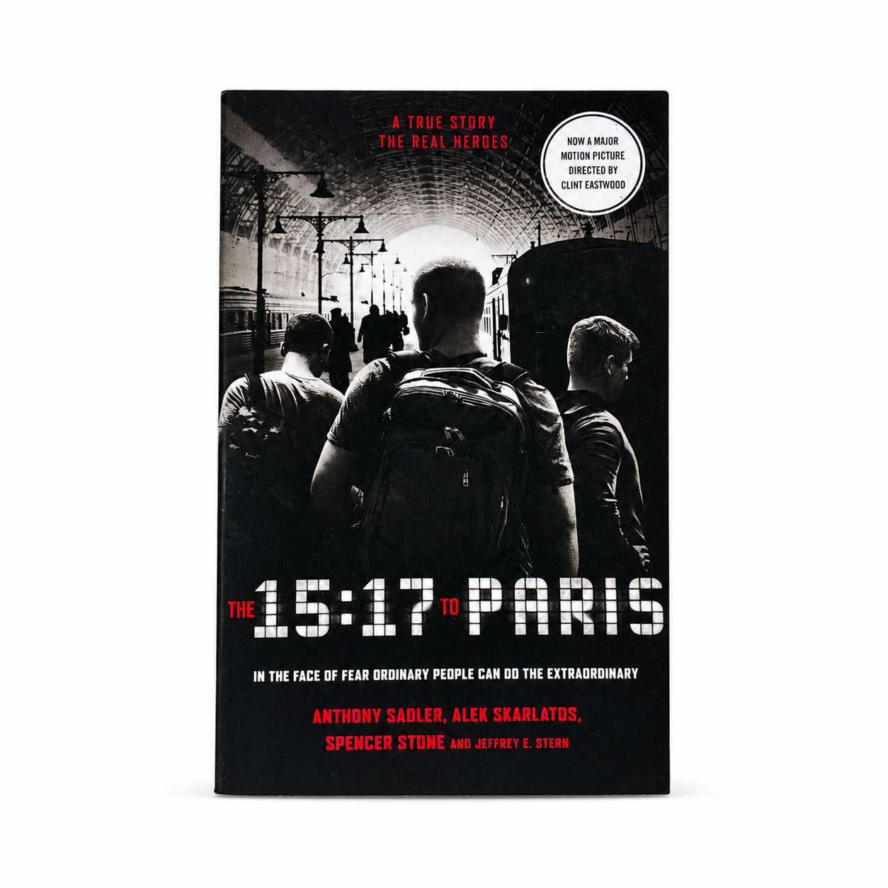 The 15:17 to Paris book cover shown upright on a light gray background. The cover is predominantly black with a grayscale photo of three people in a train station and red and white text.